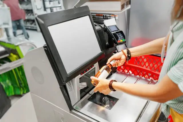 Kasa samoobsługowa: skutki automatyzacji handlu a zatrudnienie. Klient skanuje butelkę, zastanawiając się nad przyszłością pracy kasjerów.