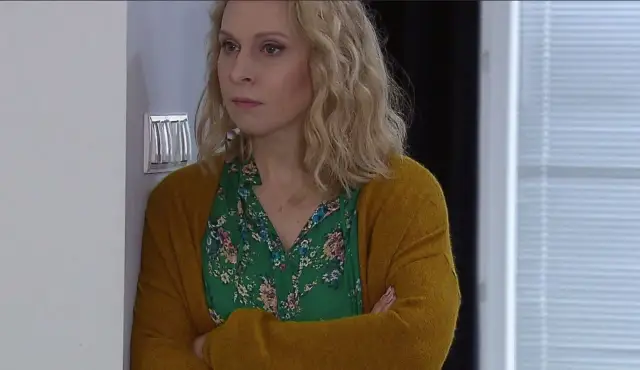 Kobieta z klanu obsada, w zielonej bluzce w kwiaty i musztardowym kardiganie, z rękami skrzyżowanymi na piersi.
