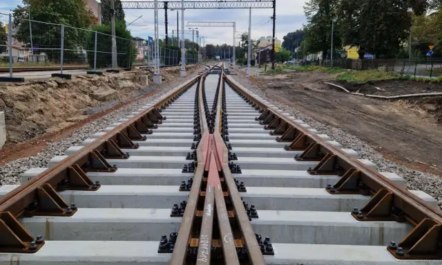 Nowe tory kolejowe, rozjazd i betonowe podkłady. W tle widać budynki i słupy trakcyjne. Akcje Torpol w budowie.