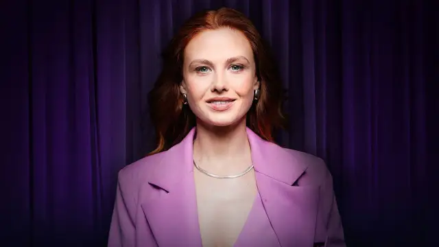 Anna Karczmarczyk w różowej marynarce, z rudymi włosami i niebieskimi oczami, uśmiecha się na tle fioletowej kurtyny. Jej filmy, seriale i programy cieszą się popularnością.