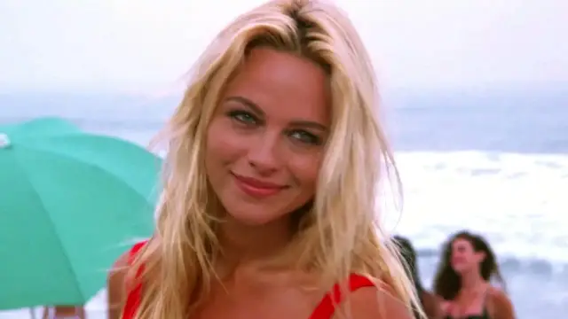 Słoneczny patrol obsada: blond ratowniczka w czerwonym stroju kąpielowym uśmiecha się na plaży, w tle ocean i parasol.