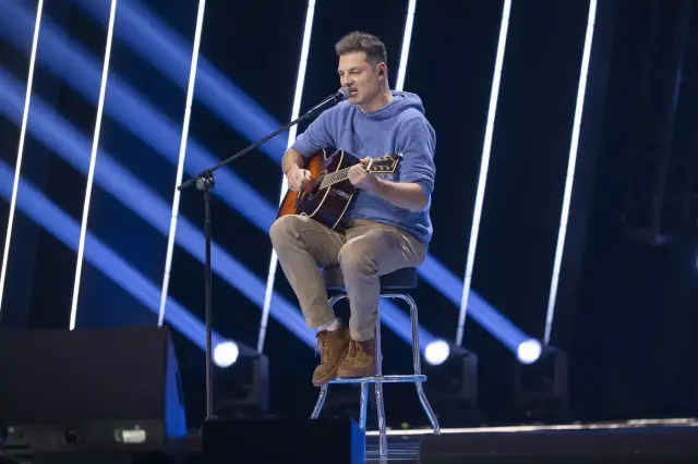 Mężczyzna siedzi na stołku, gra na gitarze i śpiewa. W tle niebieskie światła. Może to być występ z utworami o tematyce letni, chamski podryw.
