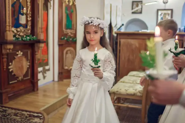 Dziewczynka w białej sukni i wianku, trzymająca świecę, przygotowuje się do komunii. Ma około 9 lat.