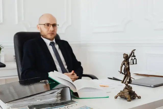 Prawnik w okularach siedzi przy biurku, obok stosu dokumentów i figury Temidy. Dowiedz się, ile kosztuje podział działki u notariusza.
