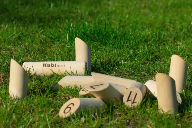 Drewniane kręgle fińskie leżą na trawie. Niektóre są ponumerowane, inne mają logo "Kubi sport".