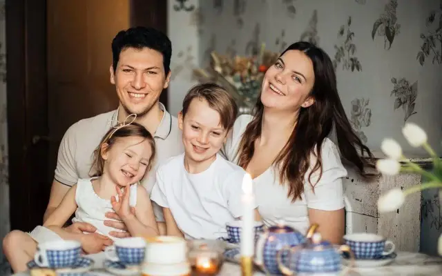 Szczęśliwa rodzina przy stole, dzieci i rodzice uśmiechają się. To obrazek, który może symbolizować radość z życia i wsparcie, jakie daje zasiłek rodzinny.