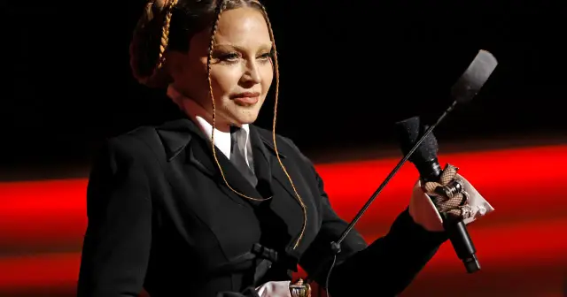 Madonna, w czarnym garniturze i z warkoczami, trzyma mikrofon i biczyk. Pytanie "czy Madonna żyje" nabiera nowego znaczenia.