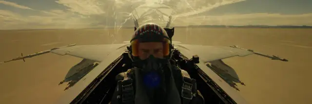 Pilot myśliwca w hełmie z napisem "Maverick" w kokpicie, lecący nad pustynnym krajobrazem.