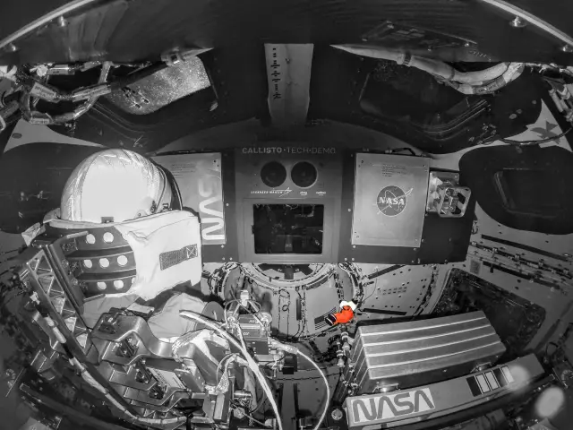 Wnętrze statku kosmicznego, widok z perspektywy astronauty. Widać hełm, panele sterowania z logo NASA i napisem "CALLISTO TECH DEMO". Mała, czerwona maskotka pingwina symbolizuje ducha misji Apollo 13.