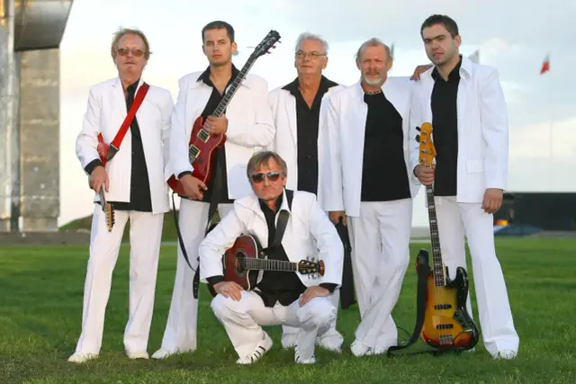 Zespół w białych garniturach, z czerwonymi gitarami i basem, gotowy do koncertu.