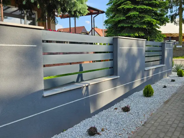 Nowoczesny płot beton architektoniczny z szarymi, poziomymi przęsłami. Ozdobne kamyczki i miniaturowe krzewy tworzą schludny ogród.