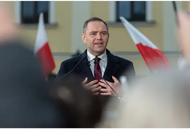 Mężczyzna w garniturze i płaszczu przemawia przy mikrofonach, za nim polskie flagi. Czy zastanawiałeś się, ile zarabia prezydent Polski?