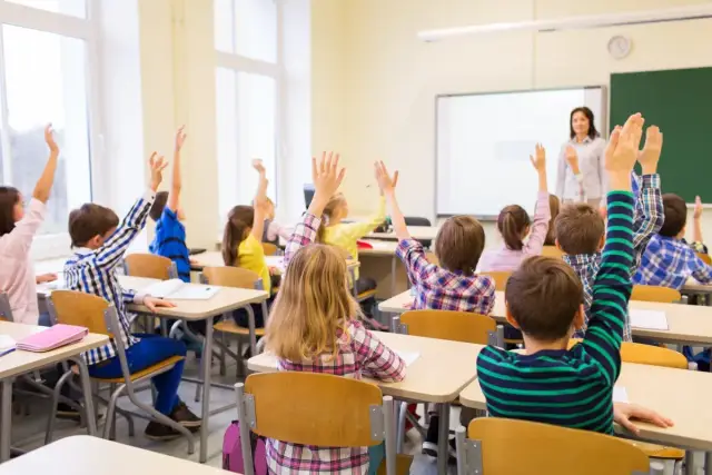 Uczniowie z podniesionymi rękami, nauczycielka wita klasę. To obraz idealny dla kogoś, kto rozważa zatrudnienie nauczyciela w trakcie studiów podyplomowych.