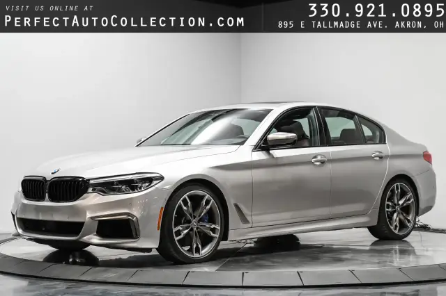 Srebrny sedan BMW m550i z czarną atrapą chłodnicy i felgami stoi na obrotowej platformie.