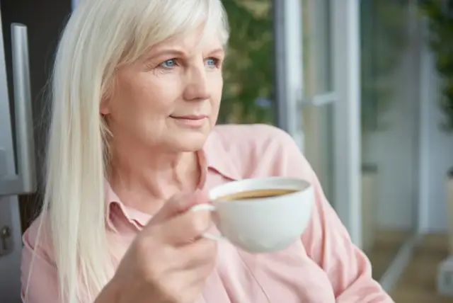 Blondynka z niebieskimi oczami popija kawę na czczo, patrząc w dal.