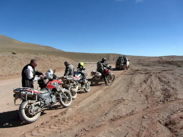 Zjazd motocyklowy w pustynnym krajobrazie. Motocykliści w pełnym rynsztunku, motocykle terenowe i samochód terenowy na piaszczystej drodze.