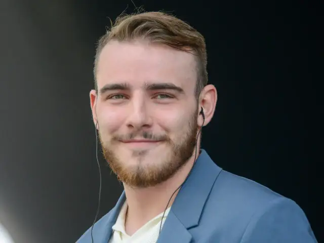 Krystian Ochman w niebieskiej marynarce, z zarostem i słuchawkami w uszach. Może właśnie rozmawia ze swoją żoną przez telefon.