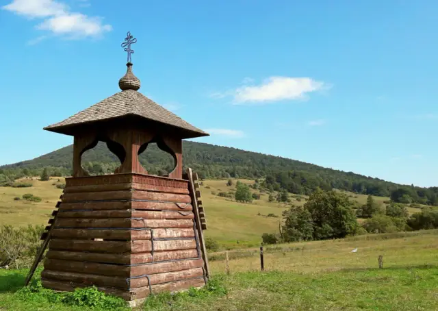 Drewniana dzwonnica z krzyżem na szczycie, otoczona zielonymi wzgórzami. To jedne z wielu regietów atrakcji.
