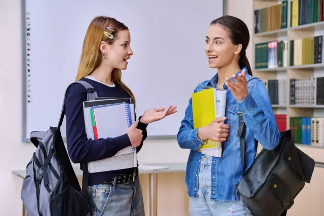 Dwie uczennice rozmawiają o tym, ile jest egzaminów zawodowych w technikum, trzymając książki i plecaki.