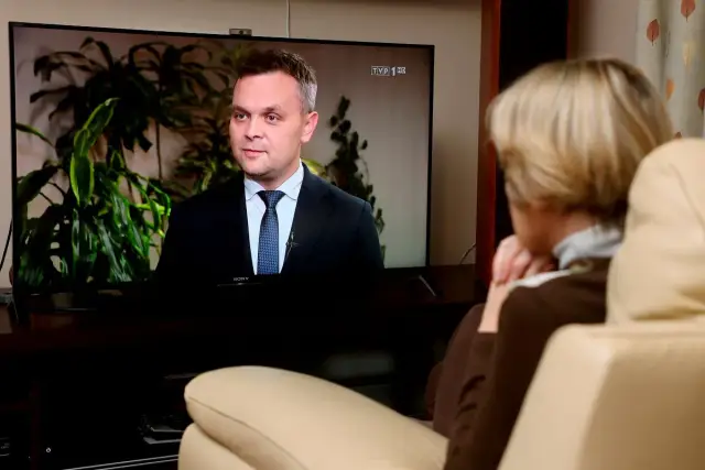 Prezes TVP w garniturze udziela wywiadu. Kobieta ogląda go z kanapy.