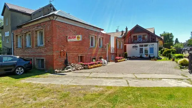 Budynek pensjonatu Aura z czerwonej cegły i białym frontem, z parkingiem i zielenią.