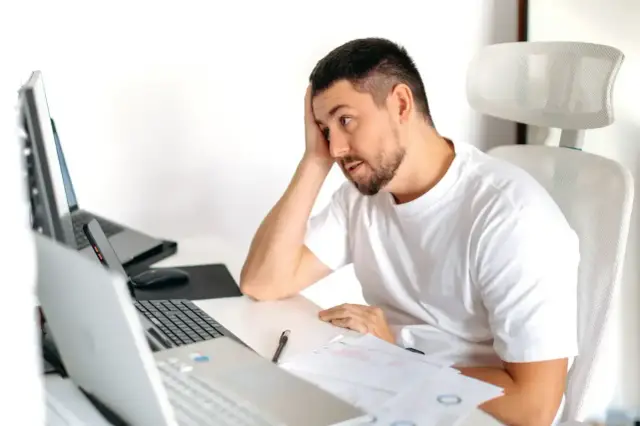 Zamyślony mężczyzna przy biurku z laptopami i dokumentami, zastanawia się, czy umowa zlecenie wlicza się do stażu pracy.