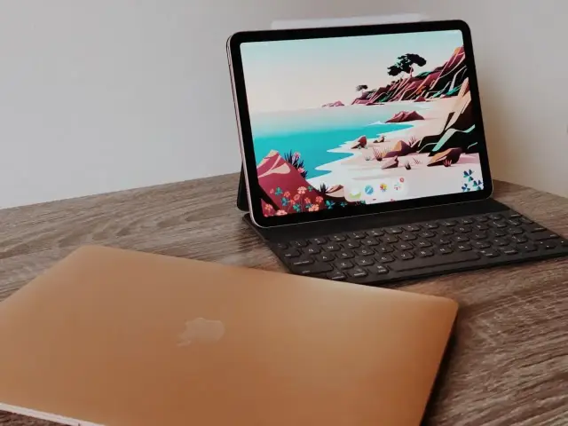 Złoty MacBook leży obok iPada z klawiaturą, który służy jako drugi ekran, wyświetlając kolorową tapetę z krajobrazem.