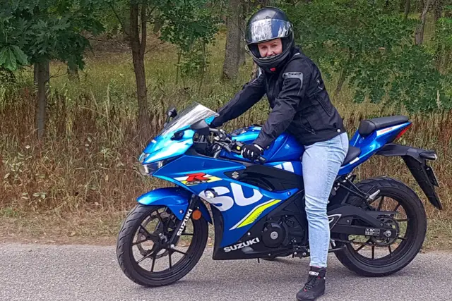 Kobieta w kasku i kurtce na niebieskim motocyklu Suzuki 125 szosowo-turystycznym, gotowa na przejażdżkę po leśnej drodze.