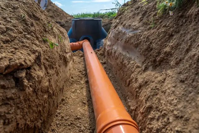 Instalacja kanalizacyjna w wykopie. Pomarańczowa rura 110 mm z minimalnym spadkiem prowadzi do studzienki.
