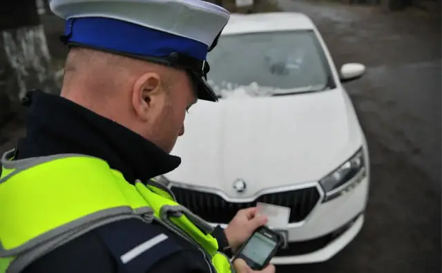 Policjant w kamizelce odblaskowej sprawdza dokumenty kierowcy przy białym samochodzie. Prowadzenie pojazdu pod kontrolą.