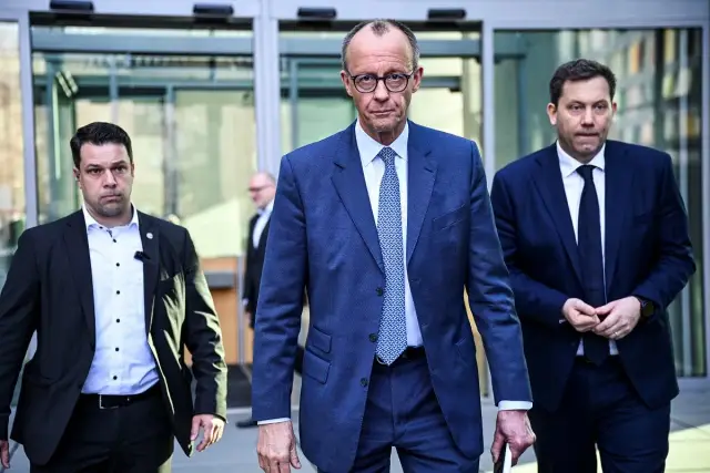Friedrich Merz, prezydent Niemiec, w niebieskim garniturze, wychodzi z budynku, otoczony ochroniarzami.