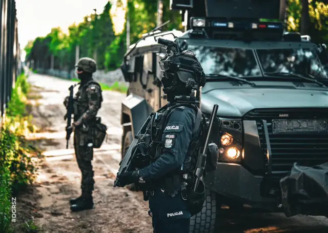 Służba mundurowa w akcji. Policjanci w pełnym rynsztunku, obok opancerzonego pojazdu, czuwają.