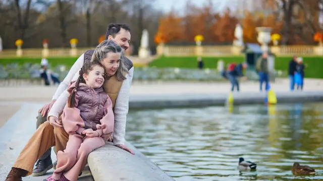 Rodzina cieszy się wspólnym spacerem nad wodą, jakby spędzali wakacje na Mazurach z dziećmi. Widok kaczek pływających w stawie dodaje uroku.