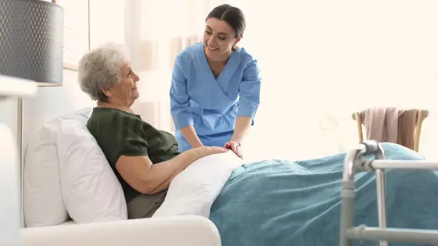Uśmiechnięta opiekunka w niebieskim uniformie wspiera starszą pacjentkę w łóżku. To obraz troski, jaki oferuje hospicjum, zapewniając komfort i wsparcie.