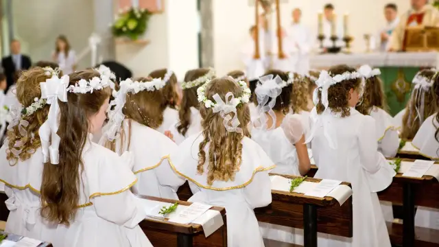 Dzieci w bieli, z wiankami na głowach, siedzą w ławkach kościoła, czekając na moment, kiedy jest komunia święta.