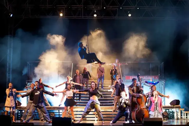 Scena musicalowa z tańczącymi i śpiewającymi ludźmi. W powietrzu wisi mężczyzna na linie, jak w musicalu.