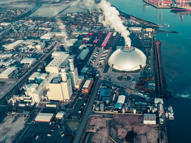 Ogromna spalarnia śmieci z charakterystyczną kopułą i kominami, z których unosi się dym. Wokół kompleks przemysłowy i port.