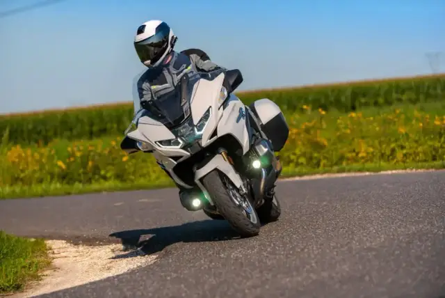 Motocyklista w kasku i stroju podróżuje na białym motocyklu BMW RT 1250 po zakręcie, z polem żółtych kwiatów w tle.