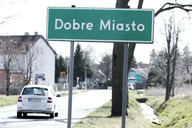 Zielony znak miejscowości "Dobre Miasto" wita podróżnych. W tle biały samochód i domy.