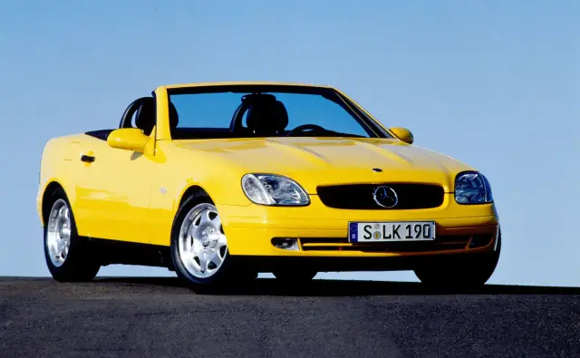 Żółty kabriolet Mercedes SLK 200 z czarnym dachem, zaparkowany na ciemnej drodze pod błękitnym niebem.