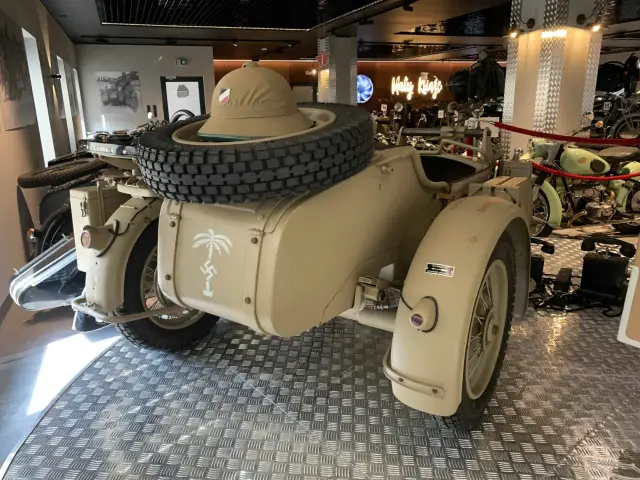 Piaskowy motocykl BMW R75 Sahara z przyczepką, kołem zapasowym i kapeluszem w tle.