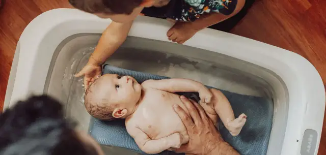 Delikatna kąpiel noworodka w wanience. Starsze dziecko z ciekawością obserwuje maluszka, a rodzice troskliwie go kąpią.