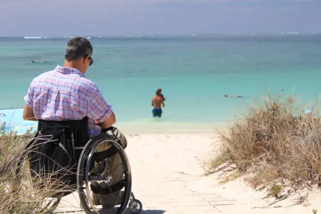 Mężczyzna z tetraplegią w wózku inwalidzkim podziwia piękny widok na turkusowe morze i plażę.