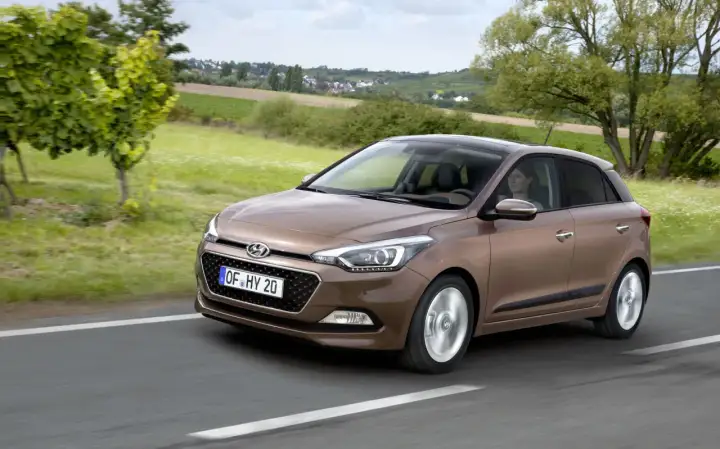 Brązowy Hyundai i20 jedzie po drodze, za nim zielone pola i domy. Sprawdź atrakcyjną hyundai i20 cena!
