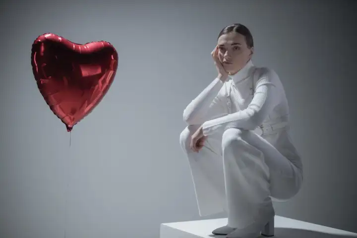 Kobieta w białym stroju siedzi na kostce, opierając głowę na dłoni. Obok niej unosi się czerwone serce-balon. Czy to symbol miłości, czy może zespół Otella?