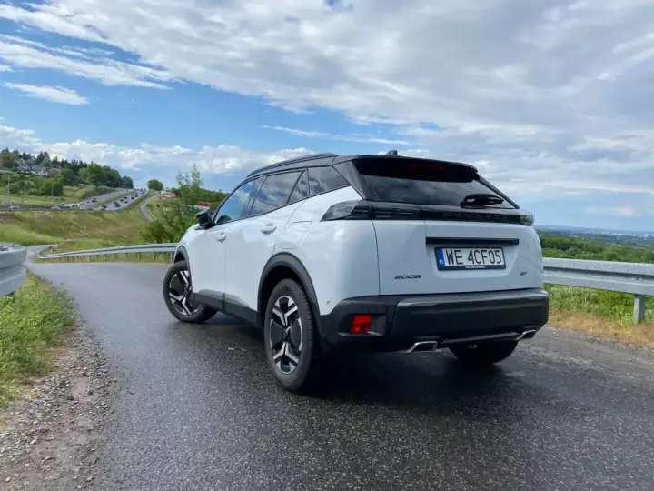 Biały Peugeot 2008 GT na mokrej drodze, z widokiem na krętą trasę i zielone wzgórza.
