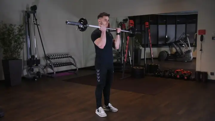 Mężczyzna wykonuje ćwiczenie barbell curl w siłowni. W tle widać sprzęt do ćwiczeń.