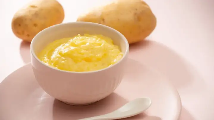 Ein Schälchen mit cremigem Kartoffelbrei, bereit für ein Baby. Zwei Kartoffeln liegen im Hintergrund.