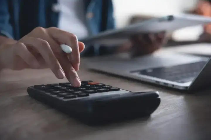 Ręka na kalkulatorze, obok laptop i dokumenty. Obliczanie, ile trzeba przepracować, by dostać 13 pensję.