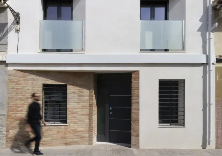 Fachada de casa con ladrillo visto y pared blanca. Una puerta oscura y ventanas con rejas. Ideas para como construir una casa barata en España.